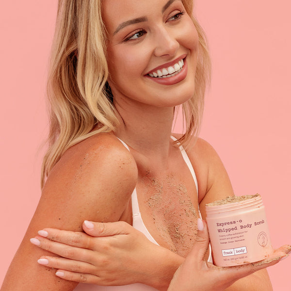 A smiling blonde woman in a white tank top holds a jar of Fresh Body Excessive Whipped Body Scrub, with scrub visible on her skin and the jar against a solid pink background.