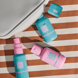 Three Dr. Brand Cosmetics travel-size skincare bottles in teal and pink, arranged on a brown and cream striped fabric surface next to a white textured pouch.
