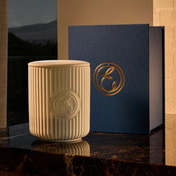 Beige textured candle with a blue box featuring a logo on a reflective surface.