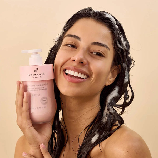 Woman with shampoo on her hair holding a pink shampoo bottle against a beige background