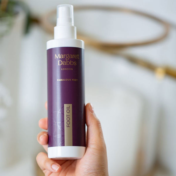 Hand holding a bottle of Margaret Dabbs London foot oil with a blurred background