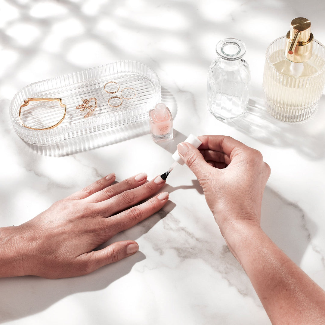 Person applying nail polish with a tray of jewelry and perfume bottles in the background.