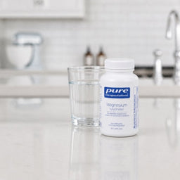 A bottle of Pure Encapsulations Magnesium Glycinate next to a glass of water on a light marble countertop in a blurred kitchen setting.