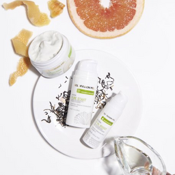 Three white skincare products from the brand GELMAGGINI are arranged on a white plate with grapefruit peel and a sliced grapefruit, alongside a jar of white cream. A hand holding a small glass pitcher is visible in the bottom right corner, set against a clean white background.